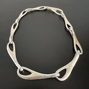 Puede incluir: Un collar de metal plateado compuesto por varios eslabones interconectados en forma de gota alargada. Cada eslabón presenta una forma orgánica, lisa y curva que se conecta con el siguiente, creando un diseño de cadena circular y fluido.