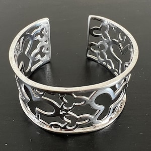 Könnte beinhalten: Ein breiter Armreif aus Silber mit einem komplizierten, durchbrochenen Ausschnittmuster aus sich wiederholenden kleeblattartigen Formen. Das Metall hat eine glatte, polierte Oberfläche und das Design umschließt das Handgelenk mit einer breiten Öffnung.