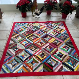 May include: A handmade quilt with a vibrant patchwork design. The quilt features a red border and a central pattern composed of colorful fabric strips arranged in diagonal lines, creating a diamond-like effect. The quilt is a mix of various colors and patterns.