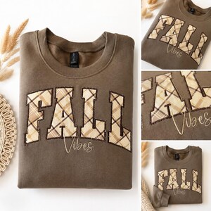 Puede incluir: Una sudadera marrón de cuello redondo con la palabra FALL en letras grandes de estilo bloque con un patrón de cuadros beige y marrón. La palabra Vibes está bordada en una fuente cursiva debajo de las letras a cuadros en la parte delantera.