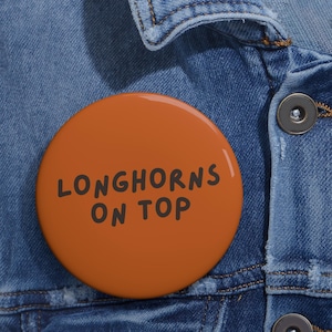 May include: A round pinback button with a burnt orange background and the black text "LONGHORNS ON TOP" printed in a simple, sans-serif font. The button is attached to a blue denim fabric surface with visible stitching and a dark metal button.