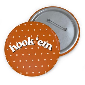 May include: A round pinback button featuring a burnt orange background with a white polka dot pattern. The text "hook 'em" is printed in white with a black drop shadow across the center. The back of the button is silver with a metal pin closure.