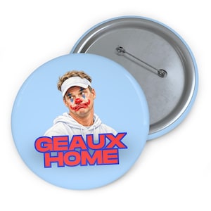 May include: A round pin-back button with a light blue background featuring an illustration of a person wearing a white visor and hoodie with clown makeup, including a red nose and blue accents. The text GEAUX HOME is printed in red and blue.