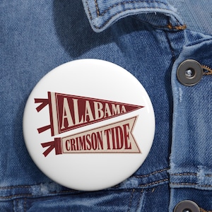 May include: A round white pinback button featuring two pennant-style graphics. The top pennant is maroon with the word ALABAMA in white, and the bottom pennant is cream with the words CRIMSON TIDE in maroon lettering, both with red ties.