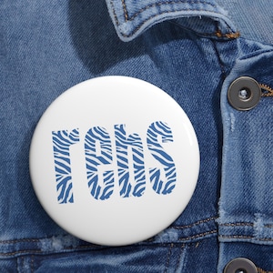 Puede incluir: Un botón circular blanco con el texto "rebs" en una fuente negrita sin remates. Las letras están rellenas con un patrón de rayas de cebra en azul y blanco. El botón está sujeto a una tela de mezclilla azul con botones metálicos visibles.