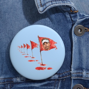 May include: A round pinback button with a light blue background featuring a series of red golf flags receding into the distance. The largest flag in the foreground displays a printed illustration of a person wearing a white visor with a neutral expression.