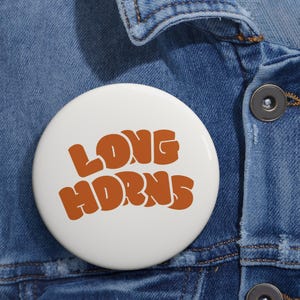 May include: A circular pinback button with a white background and the text LONG HORNS printed in a bold, wavy, burnt orange font. The button is attached to a blue denim fabric material with visible stitching and a dark metallic button detail.