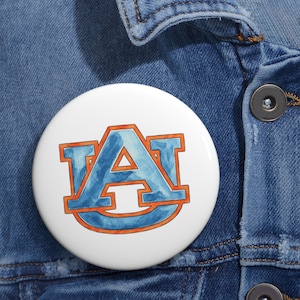 May include: A round pinback button featuring a white background with a stylized blue watercolor letter A logo outlined in orange. The letter A has a distinct serif design and is centered on the button, which is attached to a blue denim fabric.