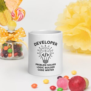Peut inclure: Un mug en céramique blanche orné d'un graphique noir représentant une ampoule avec des crochets de code à l'intérieur. Le texte indique DEVELOPER, PROBLEM SOLVER, LOGIC BUILDER, CODE WRITER. Le mug est entouré de bonbons et d'une fleur en papier jaune.