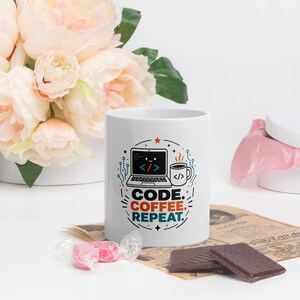 May include: A white ceramic mug featuring a graphic of a laptop with a smiling face and a coffee cup. The text reads CODE. COFFEE. REPEAT. in black, orange, and teal colours, surrounded by small stars and circuit board line patterns.