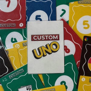 Peut inclure: Une collection de cartes à jouer UNO personnalisées comprenant une carte blanche avec le texte CUSTOM UNO au centre, entourée de diverses cartes colorées en rouge, bleu, vert, jaune et noir, affichant chacune des chiffres et le mot CUSTOM.