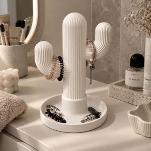 May include: A white, ribbed, cactus-shaped jewellery organiser with a circular base tray. The central column and two side arms hold various spiral hair ties and a beaded bracelet, while two patterned hair clips rest on the base tray.