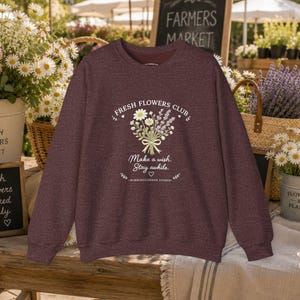 May include: A maroon crewneck sweatshirt featuring a graphic of a bouquet of daisies and lavender tied with a ribbon. The text reads "FRESH FLOWERS CLUB," "Make a wish. Stay awhile.," and "WISHINGGARDEN STUDIO" with small heart and leaf accents.