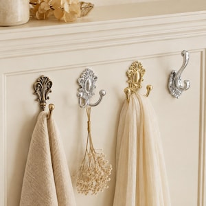 Può includere: Quattro ganci decorativi da parete montati su un pannello color crema. I ganci presentano design in metallo decorato con finiture in ottone antico, argento e oro lucido. Un asciugamano beige, un mazzo di fiori secchi e una sciarpa crema vi sono appesi.