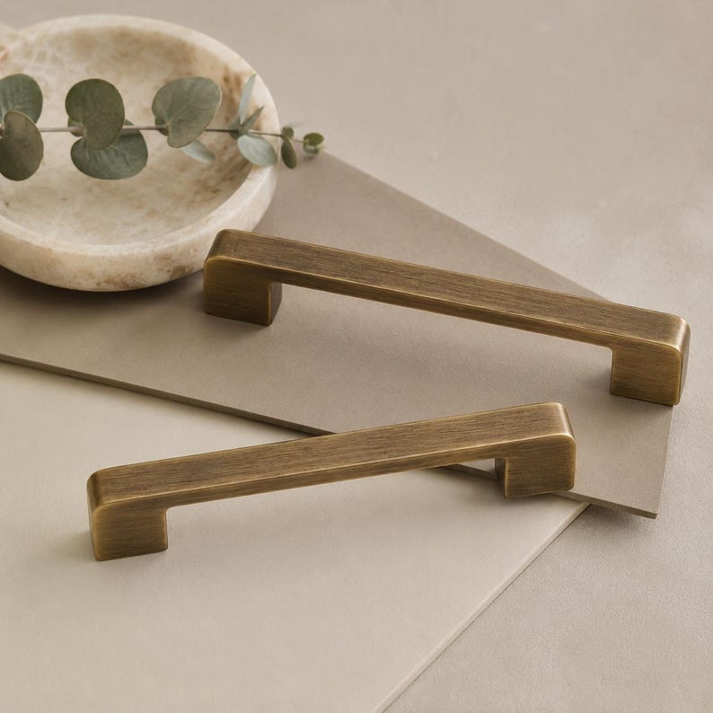 Op de afbeelding: Twee rechthoekige kastgrepen met een geborstelde antiek messing afwerking. Elke handgreep heeft een recht stangontwerp met afgeronde randen en twee bevestigingspunten aan de uiteinden, met een metalen textuur en een warme goudkleur.