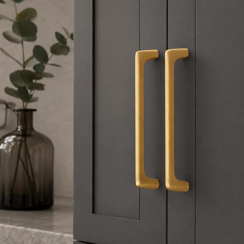 May include: Two vertical, brushed gold-toned metal cabinet pulls with a rectangular profile and rounded edges, mounted onto a dark charcoal gray cabinet door with a recessed panel design, positioned next to a dark glass vase with green foliage.