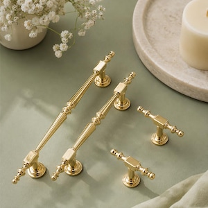 May include: A set of four polished gold-tone metal cabinet pulls and knobs featuring a slender, tapered central bar with decorative ribbed detailing and circular mounting bases. The hardware includes two long handles and two smaller T-shaped knobs.