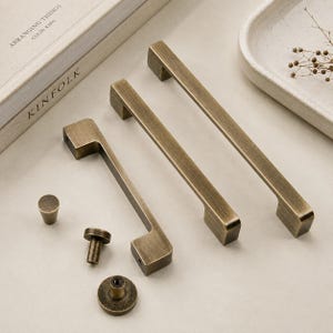 Può includere: Un set di ferramenta in ottone anticato composto da tre maniglie rettangolari per armadietti con finitura spazzolata e tre pomelli circolari abbinati. Gli elementi presentano una texture metallica usurata e sono disposti in gruppo.