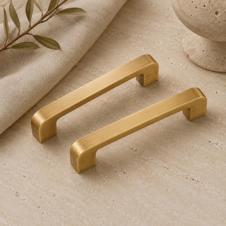 Op de afbeelding: Twee kastgrepen van geborsteld messing met een rechthoekig profiel en afgeronde randen. Deze metalen handgrepen hebben een gladde, matgouden afwerking en een eenvoudig, lineair ontwerp, wat zorgt voor een functionele grip voor meubels of kasten.