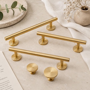 May include: Three brushed gold metal cabinet pulls with cylindrical bars and two matching round cabinet knobs. Each piece features a circular, concentric ring pattern on the mounting bases and the top surfaces of the knobs.