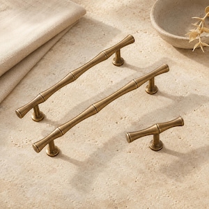 May include: Three antique brass cabinet pulls with a bamboo-inspired design. The set includes two long, slender handles with two mounting posts each and one shorter, single-post knob, all featuring a smooth, metallic finish and rounded ends.