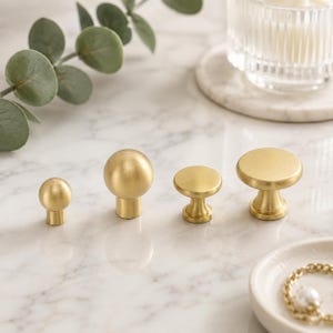 May include: Four cabinet knobs made of brushed brass metal. Two knobs feature a spherical ball shape with a cylindrical base, while the other two knobs have a flat circular top with a tapered pedestal base, all arranged in a horizontal row.