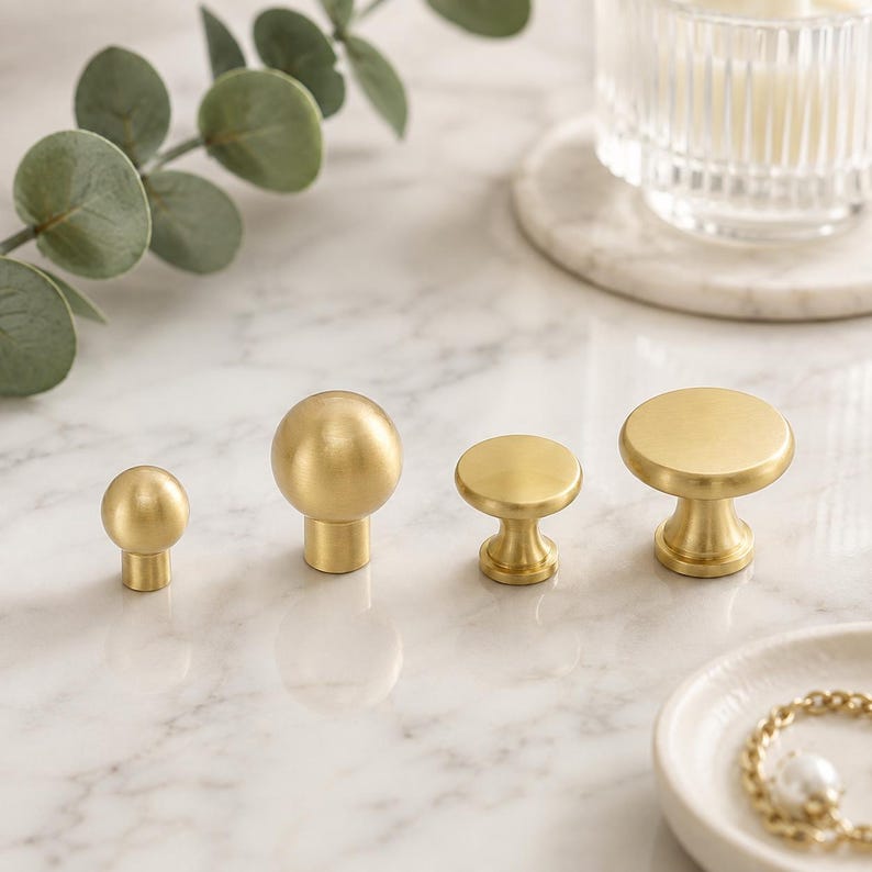 May include: Four cabinet knobs made of brushed brass metal. Two knobs feature a spherical ball shape with a cylindrical base, while the other two knobs have a flat circular top with a tapered pedestal base, all arranged in a horizontal row.
