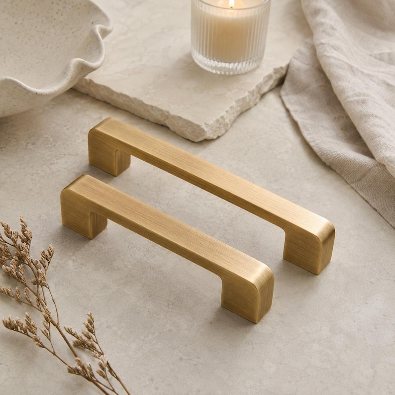 Op de afbeelding: Twee rechthoekige kastgrepen met een geborstelde gouden afwerking. Elke handgreep heeft een glad, plat bovenoppervlak met afgeronde randen en twee vierkante bevestigingspunten aan de uiteinden, met een metalen textuur en solide constructie.
