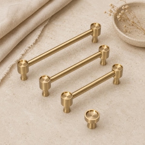 May include: A collection of three cylindrical brass cabinet pulls of varying lengths and one matching circular brass cabinet knob. Each piece features a smooth, brushed metallic finish with a simple, clean design and two sturdy mounting posts.