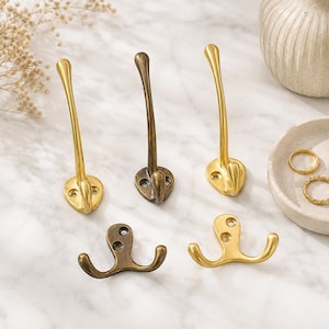 Pode incluir: Uma coleção de cinco ganchos de parede de metal com acabamentos em ouro escovado e bronze antigo. Três ganchos apresentam um design alto de ponta única com base arredondada, enquanto dois ganchos menores têm um design de ponta dupla.