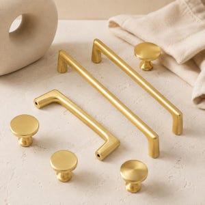 May include: A collection of brushed gold hardware including two long rectangular cabinet pulls and four round cabinet knobs. Each piece features a smooth, matte metallic finish with clean lines and a simple, functional design for furniture.