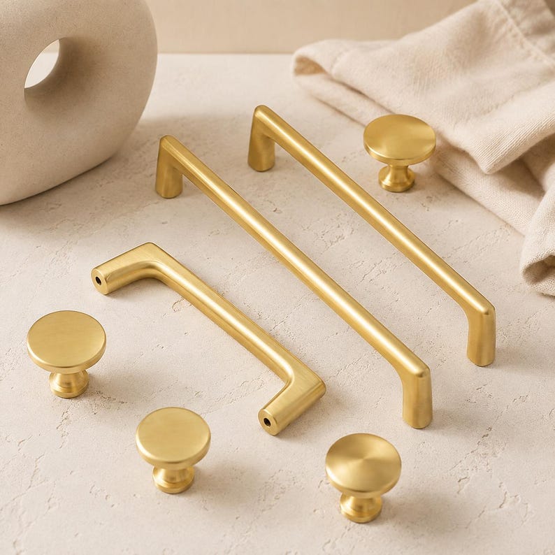 May include: A collection of brushed gold hardware including two long rectangular cabinet pulls and four round cabinet knobs. Each piece features a smooth, matte metallic finish with clean lines and a simple, functional design for furniture.
