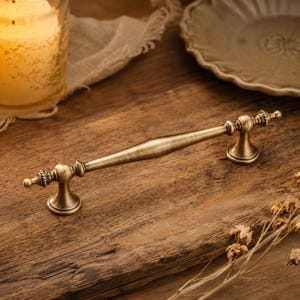May include: A long, horizontal cabinet pull handle made of brushed antique brass metal. The handle features a tapered, smooth central bar with decorative, beaded, and flared pedestal bases at each end, providing a secure mounting point.