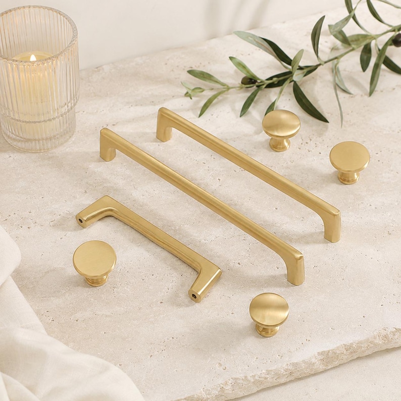 May include: A collection of brushed gold cabinet hardware including two rectangular bar pulls of different lengths and three circular knobs. The hardware features a smooth, matte metallic finish with clean lines and a simple, functional design.