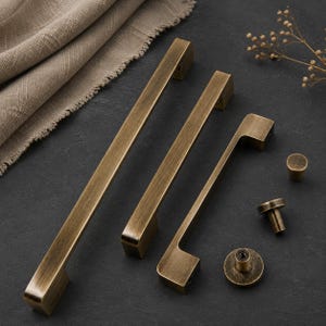 Op de afbeelding: Een set antiek messing kastbeslag bestaande uit twee lange rechthoekige ladehandgrepen, &eacute;&eacute;n kortere handgreep en drie ronde knoppen. Elk onderdeel heeft een geborstelde metalen afwerking met zichtbare donkere strepen en gladde randen.