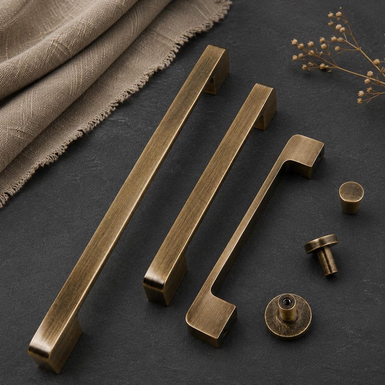 Op de afbeelding: Een set antiek messing kastbeslag bestaande uit twee lange rechthoekige ladehandgrepen, &eacute;&eacute;n kortere handgreep en drie ronde knoppen. Elk onderdeel heeft een geborstelde metalen afwerking met zichtbare donkere strepen en gladde randen.