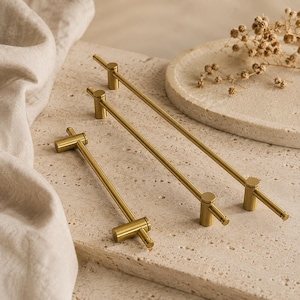 May include: Three gold-toned metal cabinet pulls of varying lengths, each featuring a slender horizontal bar supported by two cylindrical posts. The hardware has a smooth, metallic finish and a clean, linear design with small end caps on each side.