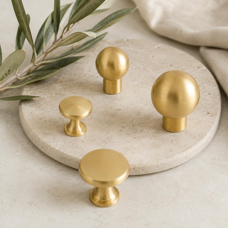 May include: A collection of four brass cabinet knobs with a brushed gold finish. The set includes two spherical knobs and two flat, circular knobs, each featuring a cylindrical base. The hardware is arranged on a circular stone surface.