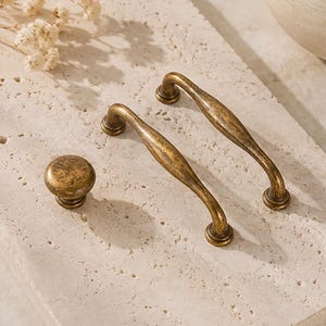 May include: Two curved metal drawer pulls and one round metal cabinet knob, all featuring an aged brass finish with a textured, mottled surface. The hardware pieces have a dark, weathered appearance with visible highlights and rounded ends.