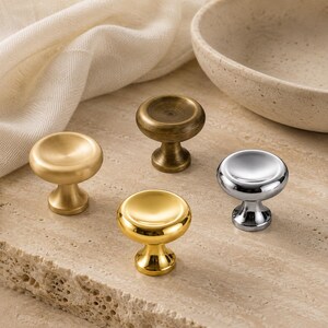 May include: Four round cabinet knobs in different metallic finishes. One knob is brushed gold, one is antique brass, one is polished gold, and one is polished chrome. Each knob features a wide, concave top surface and a tapered pedestal base.