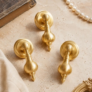 May include: Three gold-toned metal drawer pulls featuring a circular backplate and a teardrop-shaped drop handle. Each piece has a smooth, matte finish and a classic, symmetrical design with a pointed finial at the bottom of the handle.