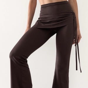 Peut inclure: Pantalon taille haute marron foncé doté d'une surjupe portefeuille intégrée. La surjupe est froncée sur la hanche droite avec de fines attaches en tissu qui pendent sur le côté. Le pantalon présente une coupe ajustée aux hanches et aux cuisses.