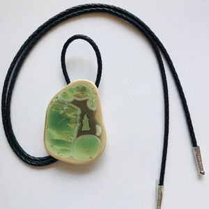 May include: A green and gold bolo tie with a unique, abstract design. The tie is made of a smooth, polished metal and has a black cord.