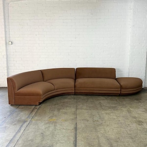 May include: A large, curved modular sofa upholstered in a solid brown fabric. The sectional piece features a low backrest, a continuous seat cushion, and a rounded end section, creating a wide, sweeping arc shape across the floor space.