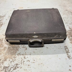 Puede incluir: Un maletín rectangular negro de carcasa rígida con un acabado exterior texturizado. El maletín cuenta con un mecanismo de cierre central de metal plateado, un asa de plástico negro y detalles de metal plateado a lo largo del borde frontal.
