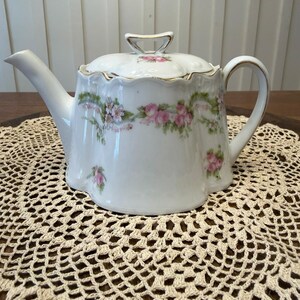 Puede incluir: Una tetera de porcelana blanca con borde festoneado, tapa a juego con asa y un delicado patrón floral de rosas rosadas y follaje verde. La tetera presenta un pico curvo y un asa, con detalles de borde dorado en la tapa y el cuerpo.
