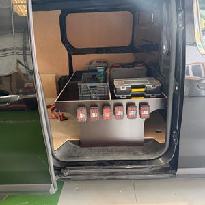 Op de afbeelding: Het interieur van een zwarte bestelwagen met een op maat gemaakte houten opbergkast met een honingraatpatroon aan de rand. De unit bevat zes rode accuhouders aan de voorzijde, met diverse gereedschapskoffers en een grijze kunststof krat bovenop.