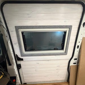 Puede incluir: Un revestimiento de pared de tablones de madera horizontales pintados de blanco instalado en el interior de un vehículo. El panel cuenta con una ventana rectangular con un marco de fieltro gris, pestillos negros y un cristal transparente.