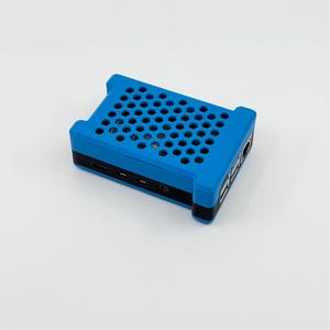 May include: A rectangular blue protective case for a single-board computer, featuring a honeycomb-patterned ventilation grid on the top surface and a black side panel with visible ports for power, data, and connectivity inputs and outputs.