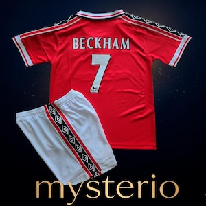 May include: A red short-sleeved football shirt with a white collar, featuring the name BECKHAM and the number 7 in white on the back, paired with white shorts. Both pieces have black and white diamond-patterned side stripes with red accents.